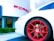 A sleek white sports car with vibrant red wheels, parked in front of a luxury car dealership. The dealership building is modern with a minimalist design, and the brand logo is prominently displayed in bold red letters.