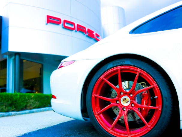 A sleek white sports car with vibrant red wheels, parked in front of a luxury car dealership. The dealership building is modern with a minimalist design, and the brand logo is prominently displayed in bold red letters.