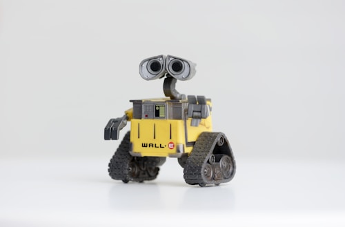 A small robot with large binocular-style eyes, tank-like treads, and a yellow cubical body, standing on a plain white surface. The robot has a distinctively cute and curious appearance.