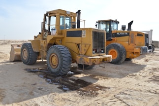 Two large yellow construction vehicles parked on a sandy surface, with clear blue skies in the background. The vehicles have large wheels and show signs of use, with dirt and wear on their surfaces.