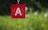 A red square keychain with a white letter 'A' in the center, surrounded by small orange rays indicating emphasis or excitement. The background is a blurred greenery, providing a contrast to the bold colors of the keychain.