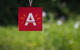 A red square keychain with a white letter 'A' in the center, surrounded by small orange rays indicating emphasis or excitement. The background is a blurred greenery, providing a contrast to the bold colors of the keychain.