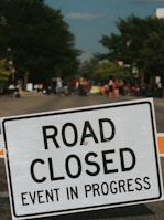 A road closure sign with the words 'ROAD CLOSED EVENT IN PROGRESS' is prominently displayed in the foreground. In the background, a blurred scene of a street with people gathered, possibly indicating a public event or festival. Trees line the sides of the road, contributing to a sense of an outdoor setting.