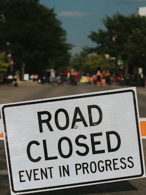 A road closure sign with the words 'ROAD CLOSED EVENT IN PROGRESS' is prominently displayed in the foreground. In the background, a blurred scene of a street with people gathered, possibly indicating a public event or festival. Trees line the sides of the road, contributing to a sense of an outdoor setting.
