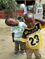 Two young boys stand on a sandy surface with a casual, playful posture. One boy wears a green and white striped polo shirt with beige pants and blue flip-flops, while the other is in a black and yellow shirt featuring the number 23 and khaki pants. In the background, a person wearing white and blue is near a food or drink stall, with tropical foliage and a brick wall adding to the beachside atmosphere.
