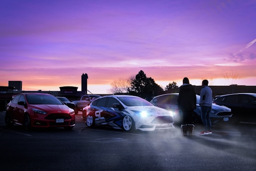 A group of modified cars is parked in a parking lot during sunset, with two people standing and conversing near the vehicles. The sky is a mix of purple and pink hues, creating a vibrant backdrop. The scene is illuminated by the headlights of one of the cars, and smoke or mist surrounds the area, adding a dramatic effect.