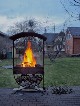 A fire pit is actively burning with bright orange flames amid a suburban backyard. The pit is made of metal, with a curved roof and storage below holding logs. It rests on a brick patio, with additional wood placed nearby. The background consists of grass, trees with bare branches, and neighboring houses under a clear blue sky.