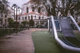 A play area features a modern slide structure with metal frames and a soft green surface. The surrounding area is empty, creating a peaceful yet somewhat deserted atmosphere. Tall trees and ornate stone architecture, with arched windows and red rooftops, dominate the background, suggesting a historic or religious building nearby the playground. Benches line the path leading to the slide, and palm trees add to the urban setting.