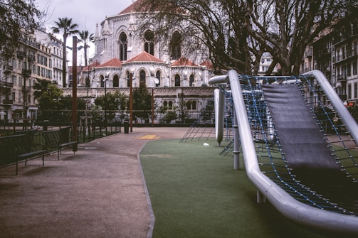 A play area features a modern slide structure with metal frames and a soft green surface. The surrounding area is empty, creating a peaceful yet somewhat deserted atmosphere. Tall trees and ornate stone architecture, with arched windows and red rooftops, dominate the background, suggesting a historic or religious building nearby the playground. Benches line the path leading to the slide, and palm trees add to the urban setting.