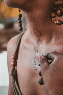 A close-up of a person's neck adorned with a large, intricately designed earring and beads hanging from the earlobe. The person wears a sleeveless garment, and a hand with fingers stained in dark pigment appears to be applying or drawing a delicate, symmetrical pattern on the skin.
