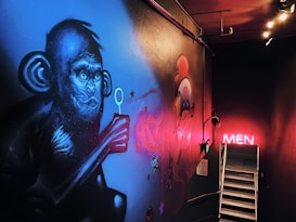 A mural featuring a stylized blue monkey holding a bubble wand covers the wall. Various artistic elements like flowers and abstract shapes surround the monkey. The scene is dimly lit with a neon pink 'MEN' sign illuminating a staircase leading downward. The vibrant artwork contrasts with the darker, shadowy environment.