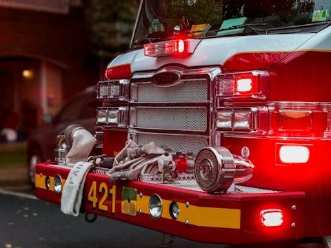 The front of a fire truck is prominently featured, showcasing its bright red color and shiny chrome details. Numerous lights are illuminated, casting a glowing and attention-grabbing effect. Coiled fire hoses and equipment are securely fastened to the truck's bumper, with the number 421 clearly visible.
