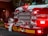 The front of a fire truck is prominently featured, showcasing its bright red color and shiny chrome details. Numerous lights are illuminated, casting a glowing and attention-grabbing effect. Coiled fire hoses and equipment are securely fastened to the truck's bumper, with the number 421 clearly visible.