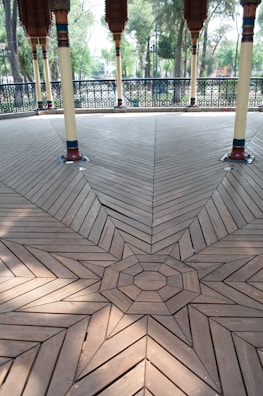 A beautifully arranged geometric wooden floor design featuring intricate patterns leading to a central octagonal shape. The wood has a light brown finish with visible grain, and the floor is supported by decorative columns with colorful accents. In the background, there are metal railings with detailed patterns, and a few trees and greenery are visible.