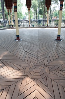 A beautifully arranged geometric wooden floor design featuring intricate patterns leading to a central octagonal shape. The wood has a light brown finish with visible grain, and the floor is supported by decorative columns with colorful accents. In the background, there are metal railings with detailed patterns, and a few trees and greenery are visible.