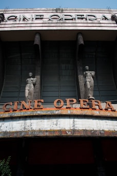 An old theater facade with large, bold letters spelling 'CINE OPERA' at the top. The structure features two statues flanking the entrance, each on a column. The building shows visible signs of wear, with peeling paint and weathering evident on the marquee. The architectural style appears to be from an earlier era, suggesting a historical significance.