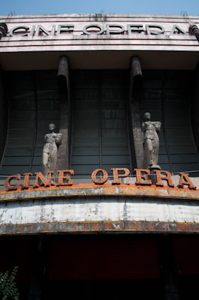 An old theater facade with large, bold letters spelling 'CINE OPERA' at the top. The structure features two statues flanking the entrance, each on a column. The building shows visible signs of wear, with peeling paint and weathering evident on the marquee. The architectural style appears to be from an earlier era, suggesting a historical significance.