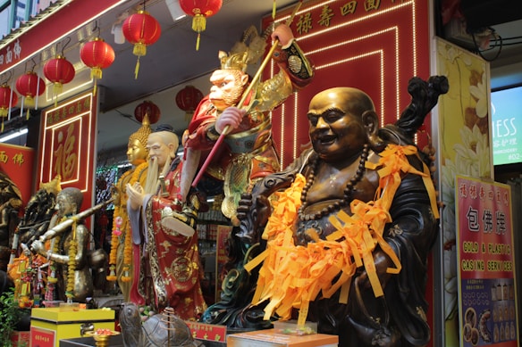 A vibrant display featuring several ornate statues of deities and figures in a shop or temple setting, adorned with gold and red elements. Bright orange ribbons and red lanterns are part of the decorative scene, creating an elaborate and festive atmosphere.