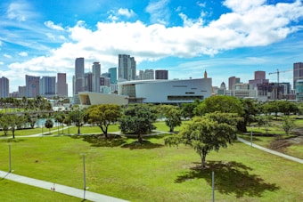 A vibrant cityscape with modern skyscrapers in the background, surrounding a contemporary architectural building. In the foreground, a large, lush green park featuring scattered trees and walking paths under a clear blue sky with scattered clouds.