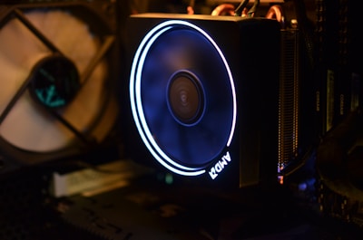A computer cooling system featuring an illuminated AMD fan with a glowing ring. The surrounding area includes other computer components such as RAM and a fan in the background. The interior is dark, highlighting the bright LED light of the fan.