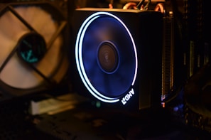 A computer cooling system featuring an illuminated AMD fan with a glowing ring. The surrounding area includes other computer components such as RAM and a fan in the background. The interior is dark, highlighting the bright LED light of the fan.