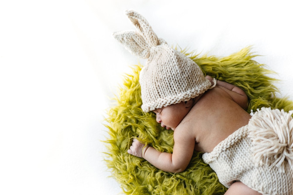 Baby in weißer Strickmütze auf grünem Felltextil liegend