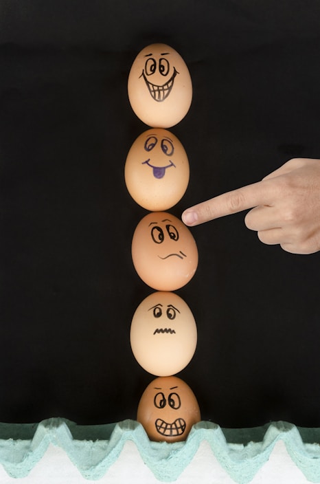 Five eggs arranged vertically, each with a different facial expression drawn on them using a black marker. From top to bottom: a wide smile, a playful tongue out, a worried face, a scared face, and an angry face. A human hand is pointing towards the middle egg. The eggs are placed against a dark background and rest on a green egg carton.