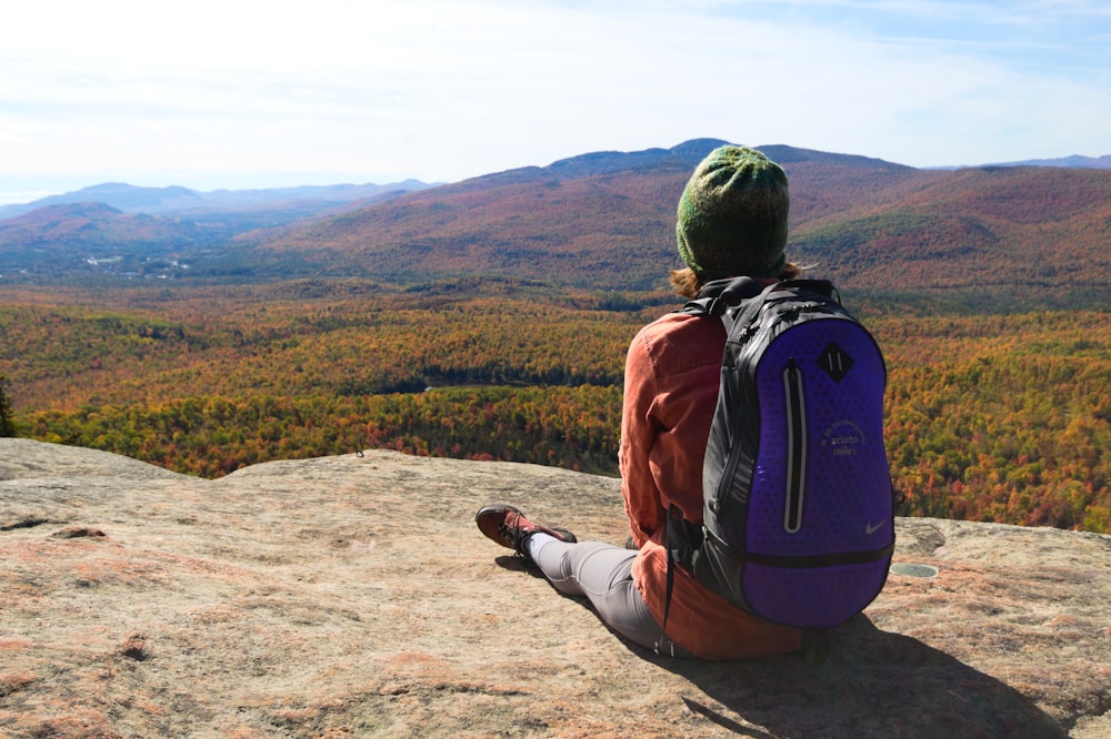 man in orange jacket and black pants sitting on brown rock during daytime