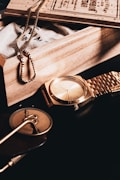 A collection of elegant accessories arranged artfully on a dark surface. A gold watch with a matching metallic band sits prominently. Next to it, there's a gold-colored chain draped over a textured cloth bag within a wooden box. A round, decorative medallion and some cards or booklets can also be seen.