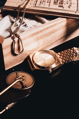 A collection of elegant accessories arranged artfully on a dark surface. A gold watch with a matching metallic band sits prominently. Next to it, there's a gold-colored chain draped over a textured cloth bag within a wooden box. A round, decorative medallion and some cards or booklets can also be seen.