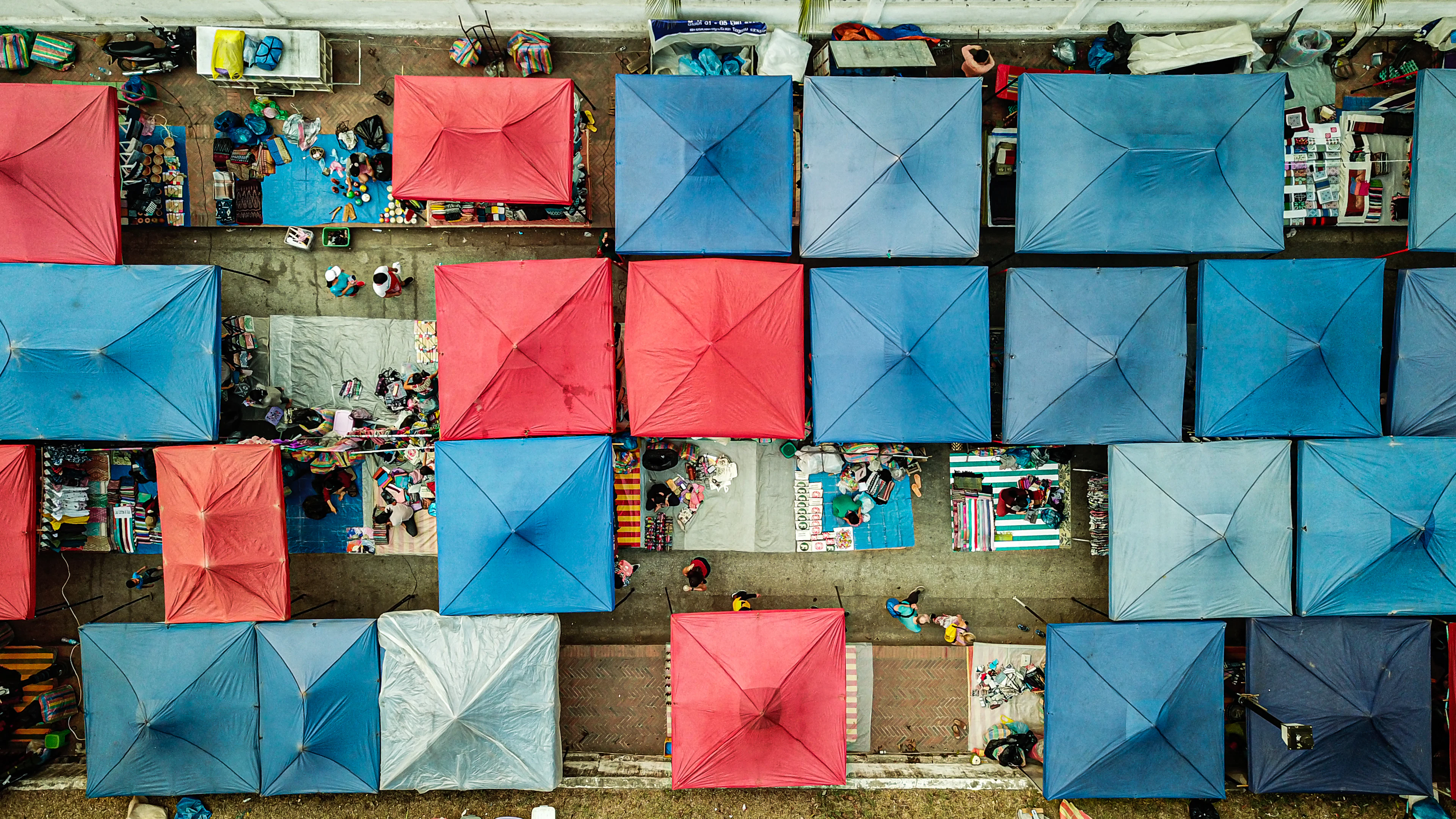 Luang Prabang's night market seen from directly above, a grid of red and blue canvas tent roofs pitched down the street with vendors and their laid-out textiles in the gaps between them