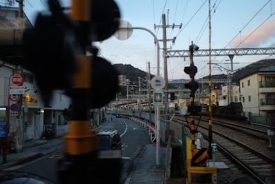 A railway crossing at an urban area with power lines, tracks, and signaling equipment. The street adjacent to the railway has a car and various street signs. Residential buildings and a mountainous background are visible, creating a blend of industrial and residential scenery.