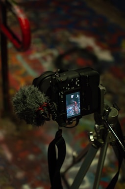 A camera mounted on a tripod with an attached microphone, capturing a scene projected on its screen. The background is a colorful, abstract surface with splashes of various hues.