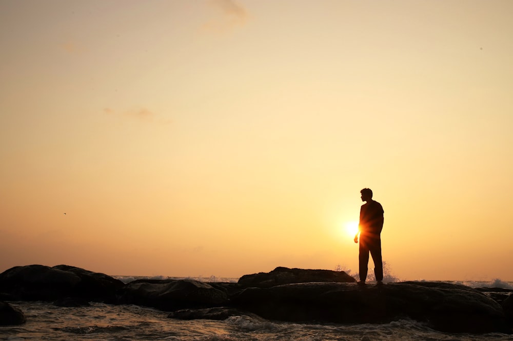 man standing on rock formation near body of water during sunset