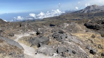 A rugged mountainous landscape with a rocky trail winding through the terrain. The image features mountains in the background under a clear blue sky with scattered clouds. The area is mostly barren with patches of dry grass and rocky outcrops dominating the scene.