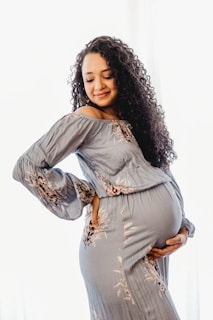 A pregnant woman wearing a floral-patterned dress holds her belly with one hand while gently resting the other on her back. She has long, curly hair and a serene, content expression on her face.
