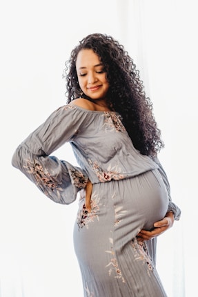A pregnant woman wearing a floral-patterned dress holds her belly with one hand while gently resting the other on her back. She has long, curly hair and a serene, content expression on her face.
