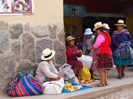 A group of women wearing traditional clothing and hats are gathered around selling and buying fruit in a market. They are sitting beside a stone wall with colorful blankets and bags. Some fruit is displayed on a cloth on the ground.