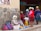 A group of women wearing traditional clothing and hats are gathered around selling and buying fruit in a market. They are sitting beside a stone wall with colorful blankets and bags. Some fruit is displayed on a cloth on the ground.