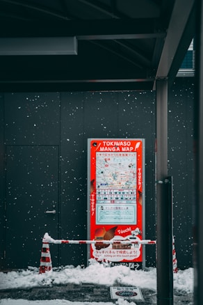 A red sign labeled 'TOKIWASO MANGA MAP' stands against a dark wall. Snow falls gently, covering the ground and creating a wintry scene. A red and white traffic cone and barrier are also visible, partially covered in snow. The scene suggests a cold, outdoor setting.