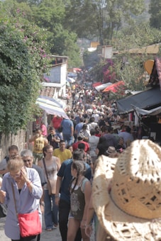 A bustling outdoor market filled with people walking along a narrow street lined with various stalls. The dense crowd features individuals of all ages, some carrying bags or wearing hats. Lush greenery and colorful flowers adorn the surrounding buildings, creating a vibrant atmosphere.