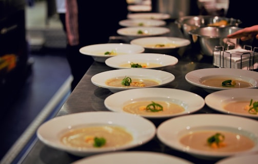 Numerous white plates are neatly arranged on a metal countertop, each containing a portion of creamy soup garnished with green herbs. The setting suggests a professional kitchen or a restaurant where the dishes are being prepared for serving. The background is slightly blurred, with bowls and ingredients visible, adding to the culinary atmosphere.