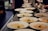 Numerous white plates are neatly arranged on a metal countertop, each containing a portion of creamy soup garnished with green herbs. The setting suggests a professional kitchen or a restaurant where the dishes are being prepared for serving. The background is slightly blurred, with bowls and ingredients visible, adding to the culinary atmosphere.