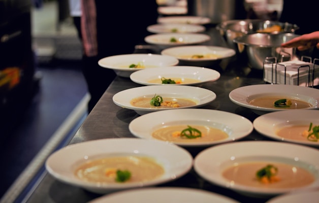 Numerous white plates are neatly arranged on a metal countertop, each containing a portion of creamy soup garnished with green herbs. The setting suggests a professional kitchen or a restaurant where the dishes are being prepared for serving. The background is slightly blurred, with bowls and ingredients visible, adding to the culinary atmosphere.