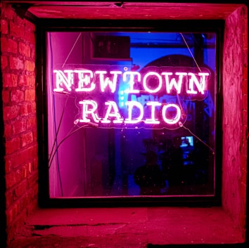 A vibrant neon sign displays the words 'Newtown Radio' in pink, bordered by a window frame. The sign is illuminated from within a dimly lit room, casting a warm, glowing reflection. On the left, the exposed brick wall adds texture. Inside the room, silhouettes are barely visible, contributing to a mysterious ambiance.