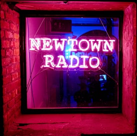 A vibrant neon sign displays the words 'Newtown Radio' in pink, bordered by a window frame. The sign is illuminated from within a dimly lit room, casting a warm, glowing reflection. On the left, the exposed brick wall adds texture. Inside the room, silhouettes are barely visible, contributing to a mysterious ambiance.