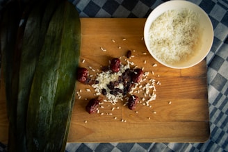 A wooden cutting board displays a small pile of uncooked white rice mixed with raisins and dates. Adjacent to the pile is a bowl filled with more white rice. On the left, large green leaves are neatly arranged. The background features a checkered blue and white tablecloth.