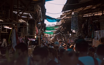 A bustling market with a narrow passageway lined with stalls on both sides. The stalls are covered with fabric canopies and display various goods, including textiles and souvenirs. The area is crowded with people, creating a dynamic and lively atmosphere.