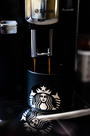 A coffee machine dispenses fresh coffee into a black cup featuring a well-known mermaid logo. The reflection of the cup and logo can be seen on the shiny surface below.