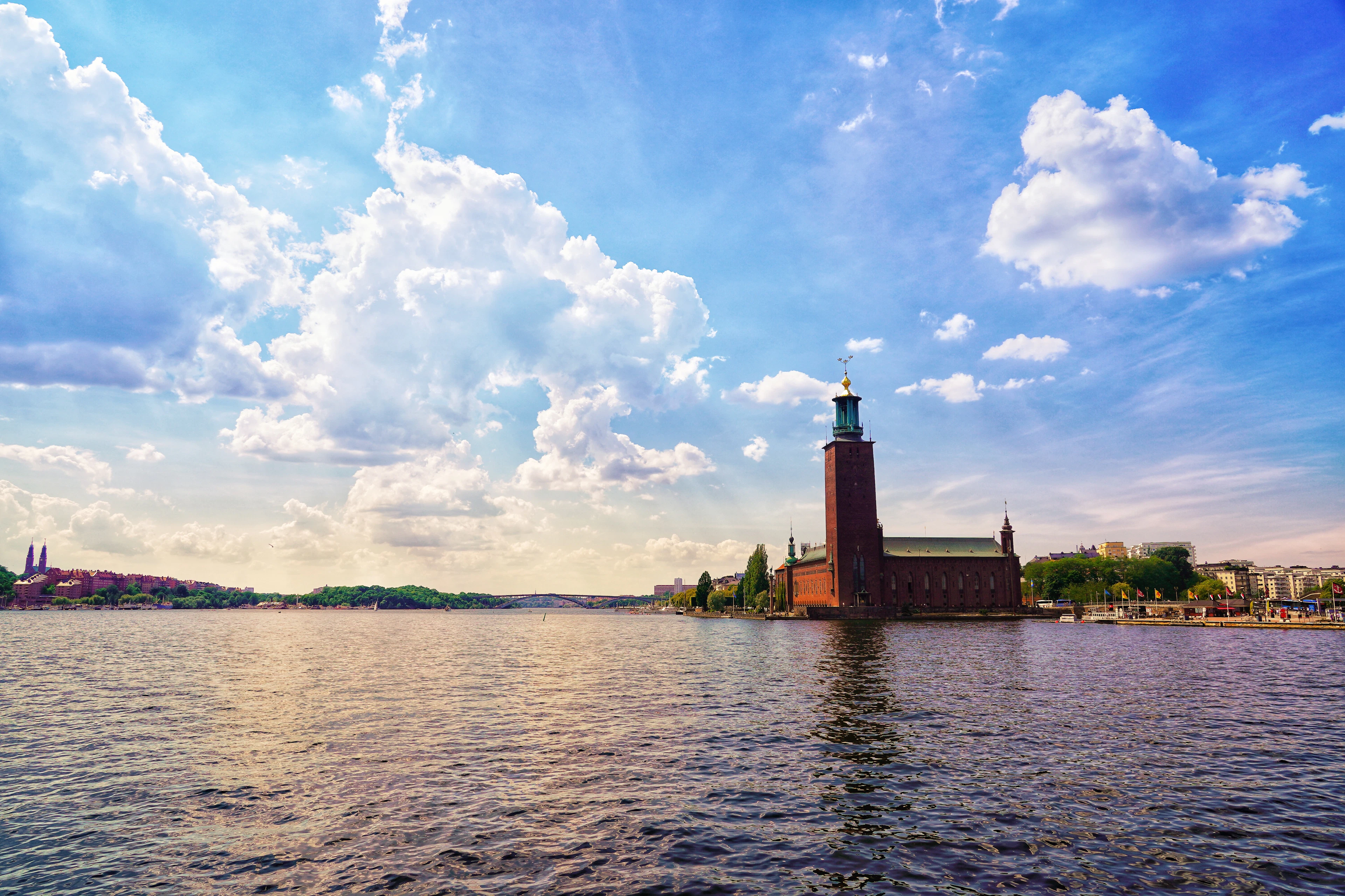 Stockholm City Hall's brick tower rises beside the water under a bright blue sky with billowing clouds.